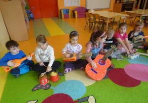 Grupa dzieci siedzi na brzegu dywanu z instrumentami muzycznymi wykonanymi z materiałów pochodzących z recyklingu.